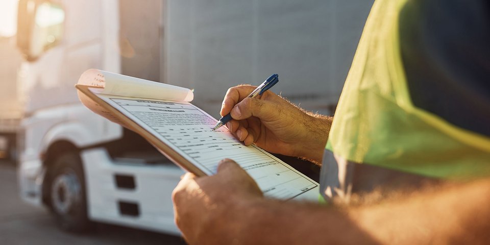 Auf dem Foto sieht man die Hände eines Mannes, der ein Klemmbrett mit mehreren Papieren hält. Das oberste Blatt ist ein Formular. Mit der rechten Hand füllt er mit einem Kugelschreiber das Formular aus. Im Hintergrund sieht man den vorderen Teil eines Lastkraftwagens mit dem Führerhaus.